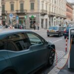 Carro elétrico sendo carregado em estação pública na Europa com veículos a gasolina ao fundo