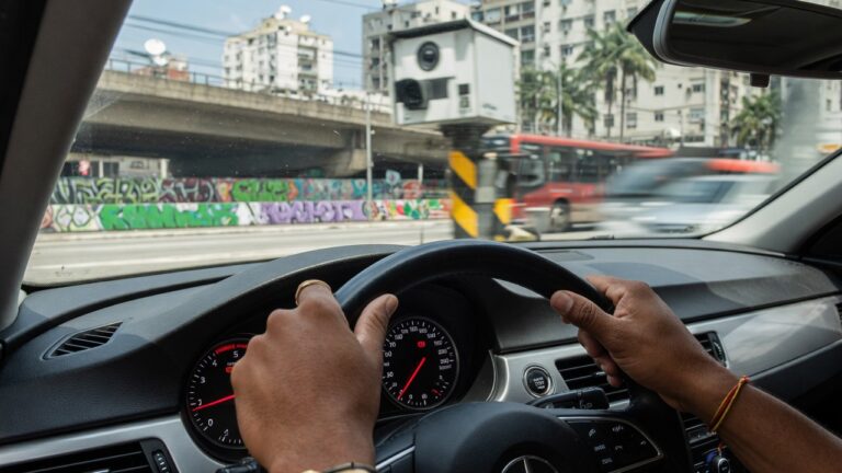 Motorista ao volante de carro moderno com painel de instrumentos visível e radar de trânsito ao fundo, representando infrações de trânsito